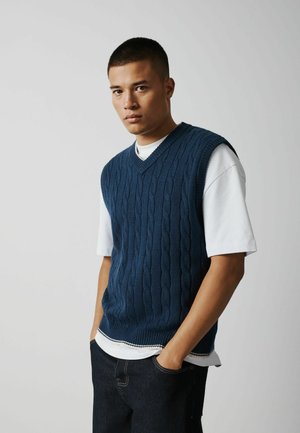 Young man wearing a navy blue cable-knit sleeveless sweater over a white t-shirt, standing with hands in pockets against a plain background.