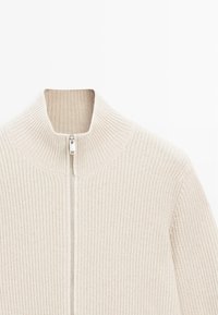 Felpa zip con colletto alto di colore crema, caratterizzata da una texture a coste e una cerniera metallica lucida, crea una silhouette strutturata.