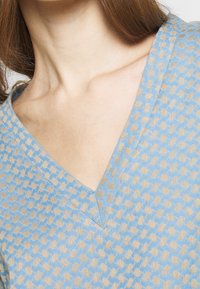 Jasnoniebieska bluzka z dekoltem w serek, z teksturowanym wzorem w beżu, wykonana z miękkiego materiału i w luźnym kroju, podkreślająca design dekoltu.