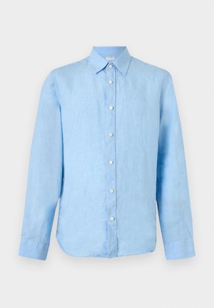 Homme debout portant une chemise bleu clair à manches longues boutonnée, un short beige, des chaussettes blanches et des baskets blanches sur un fond uni.
