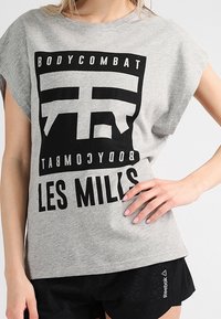 Camiseta gris de manga corta con estampado gráfico negro que dice "BODYCOMBAT" y "LES MILLS" en el frente, combinada con pantalones cortos negros que llevan el logo.