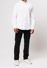 Valkoinen pitkähihainen kauluksellinen napillinen paitapusero, yhdistetty mustiin housuihin ja valkoisiin tennareihin. Päällä pieni logo rinnassa.