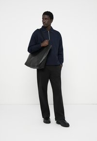 Sudadera de polo navy con cuello de cremallera, bolsa tote negra, pantalones anchos negros y zapatos negros resistentes, sobre un fondo blanco.