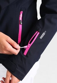 Námořnicky modrá nepromokavá bunda s růžovými kapsami na zip. Má nastavitelné manžety na suchý zip a měkký, pružný materiál pro pohodlí.