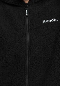 Schwarze Fleecejacke mit strukturiertem Stoff, Reißverschluss und weißem, gesticktem "Bench."-Logo auf der Brust und am Reißverschluss-Ziehgriff.