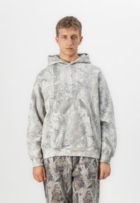 Giovane uomo che indossa un felpa con cappuccio e pantaloni in camouflage grigio e beige, in piedi di fronte su uno sfondo bianco uniforme.