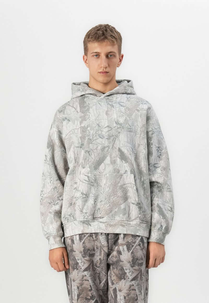 Giovane uomo che indossa un felpa con cappuccio e pantaloni in camouflage grigio e beige, in piedi di fronte su uno sfondo bianco uniforme.