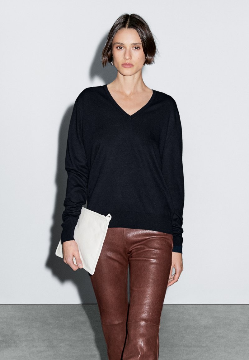 Pullover nero a V con maniche lunghe, abbinato a pantaloni in pelle marrone e una borsa a tracolla bianca. Texture lisce e design contemporaneo.