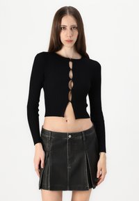 Haut court en tricot côtelé noir avec des manches longues et une fermeture à bouton en forme de trou de clé. Associé à une mini jupe en cuir noir dotée de coutures et de poches.
