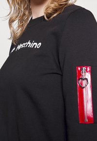 Černá mikina s bílým logem "Moschino" a červenou zipovou kapsou na rukávu, zvýrazněná zipem ve tvaru srdce.