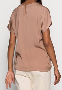 Blusa marrone a maniche corte con scollo rotondo, dotata di un piccolo dettaglio a chiave con bottone sul retro e una texture liscia.