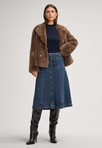 Giacca doppio petto in pelliccia sintetica marrone, maglietta blu navy a maniche lunghe a coste, gonna A-line in denim blu con bottoni e stivali neri in pelle alti.