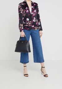 Žena v květinovém černém blejzru, modrých zkrácených džínách, černých lodičkách na vysokém podpatku, s černou koženou kabelkou s krátkými rukojeťmi.