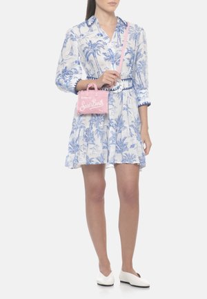 Donna che indossa un vestito bianco con stampa di palme blu, tiene una piccola borsa a tracolla rosa e indossa scarpe basse bianche.