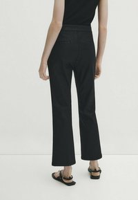 Donna che indossa pantaloni neri a gamba larga e corta, top senza maniche nero e sandali piatti neri con cinturini alla caviglia, in piedi davanti a uno sfondo bianco.