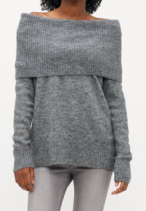 Femme portant un pull gris côtelé à épaules dénudées associé à un jean gris clair ajusté sur un fond uni.