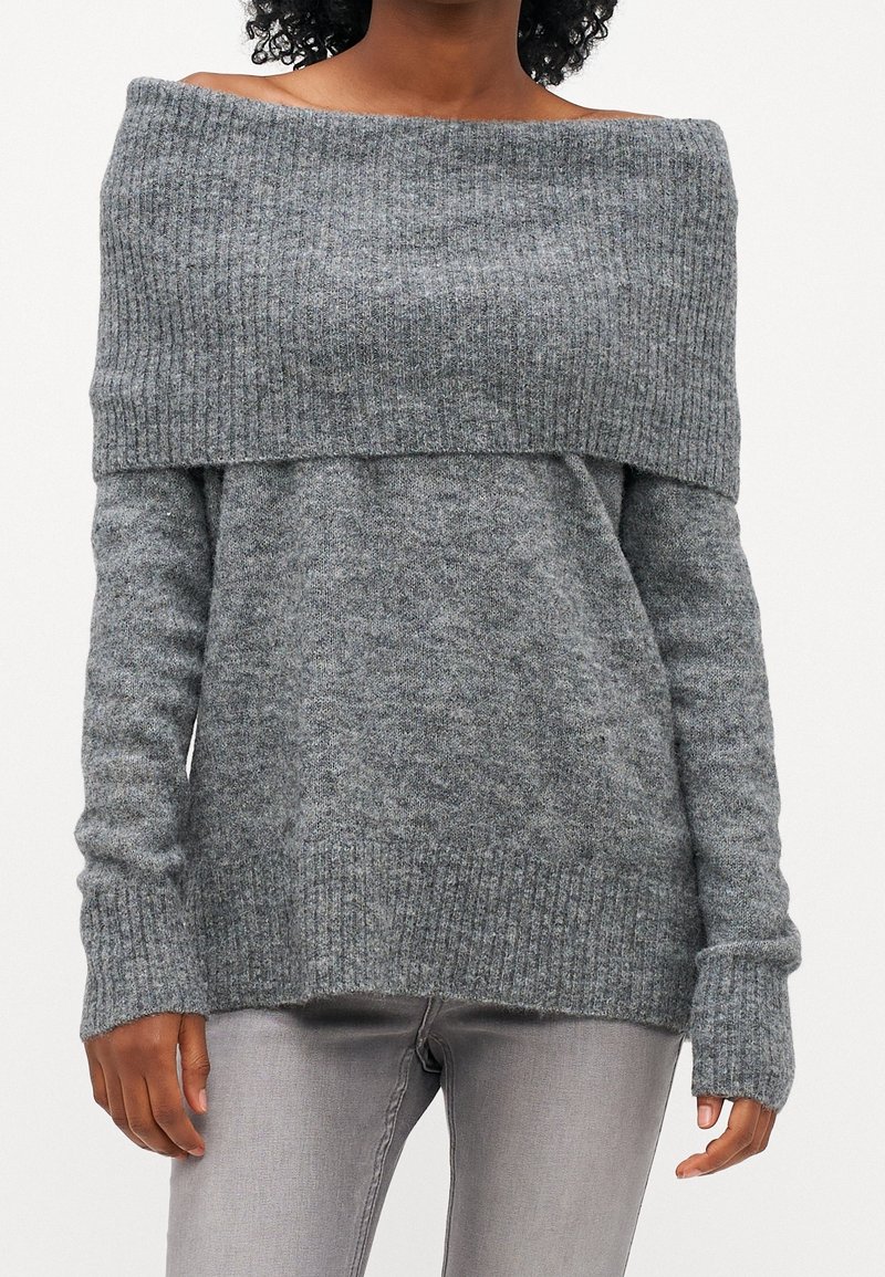 Femme portant un pull gris côtelé à épaules dénudées associé à un jean gris clair ajusté sur un fond uni.