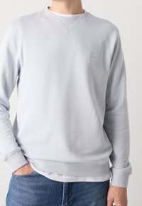 Homme portant un sweat-shirt gris clair à manches longues avec un petit logo sur la poitrine et un jean bleu, une main dans la poche sur fond uni.