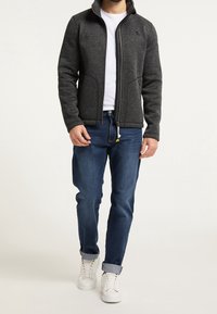 Šedá bunda na zip s vysokým límcem, texturovaná látka, dvě přední kapsy, nošená přes bílé tričko a modré džíny. Bílé tenisky s ohrnutými lemy.