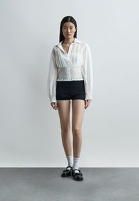 Νεαρή γυναίκα σε όρθια στάση, ντυμένη με διάφανο λευκό μπλουζάκι, μαύρο σορτς, λευκές κάλτσες και μαύρα παπούτσια, μπροστά σε απλό ανοιχτό γκρι φόντο.