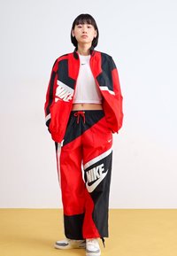 Piros és fekete Nike tréningöltöny geometrikus mintával. Cipzáras dzseki és zsinóros nadrág jellemzi, mindkettőn nagy logók láthatók.
