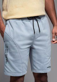 Shorts en polaire bleu clair avec une taille élastique à cordon, coutures latérales et texte imprimé noir sur la jambe gauche. Texture lisse et longueur au-dessus du genou.