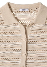 Cardigan beige et blanc en maille ajourée avec col côtelé et boutons beiges, avec une étiquette de la marque "CECIL" à l'intérieur de l'encolure.