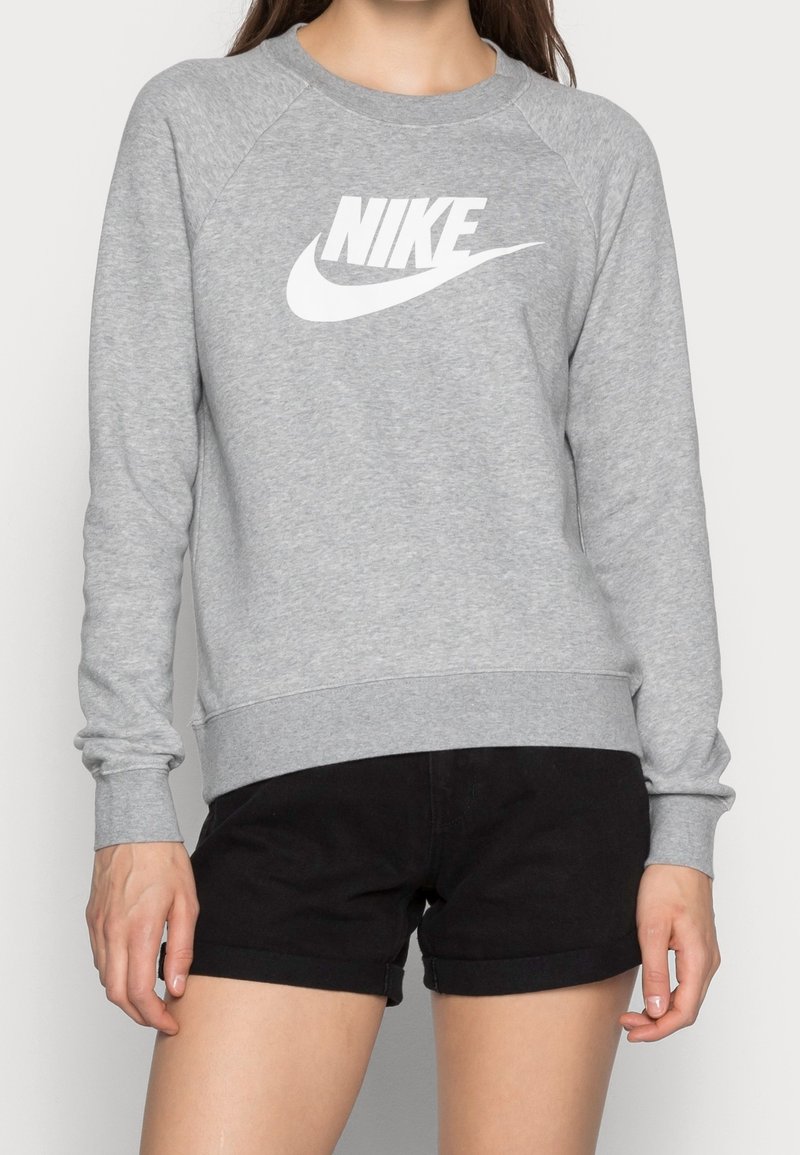 Femme portant un sweat-shirt Nike gris clair avec logo à l'avant et un short noir, debout les bras détendus le long du corps.