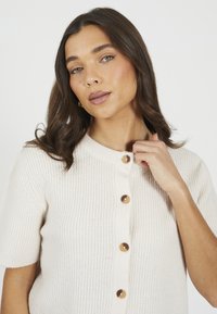 Krótki rękaw, kremowy kardigan o ryflowanej fakturze i okrągłym dekolcie, z pięcioma dużymi, błyszczącymi guzikami wzdłuż przodu.
