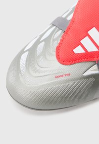 Scarpa sportiva argentata con tomaia testurizzata, accenti rossi, marchio evidente e design aerodinamico con motivi di presa visibili.