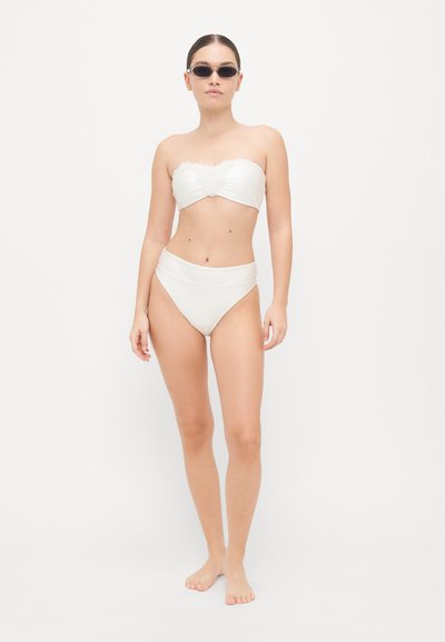 Bikini blanc sans bretelles avec un haut texturé et un bas taille haute. Modèle portant des lunettes de soleil noires, debout sur un fond uni.