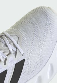 Scarpa sportiva bianca con tomaia in maglia texturizzata, lacci piatti e accenti scuri sui lati, dotata di una suola in gomma imbottita.
