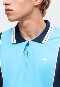Jeune personne portant une chemise bleu clair à fermeture éclair avec un col bleu foncé et blanc, tournée vers la gauche avec une expression neutre.
