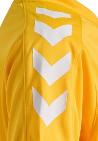 Gul sportströja med textilstruktur och vitt chevronmönster över axlarna. Har rund halsringning och lös passform.