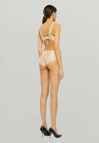 Beige bralette och högmidjade trosor i slätt tyg med en sömlös design. Modellen bär svarta högklackade skor.