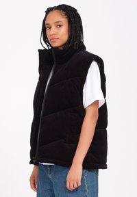 Schwarze Cord-Puffer-Weste mit hohem Kragen und Reißverschluss. Verfügt über ein gesteppte Design, seitliche Taschen und wird über einem weißen T-Shirt getragen.