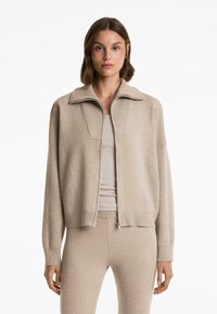 Beige stickad zip-jacka med hög krage och ribbade mudd, som har ett texturerat vävmönster och en kortad siluett.