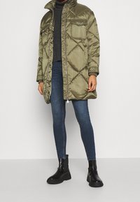 Una persona está de pie vistiendo un abrigo de plumas verde oliva de gran tamaño, un suéter gris, jeans ajustados azules y botas negras hasta el tobillo, frente a un fondo sencillo.