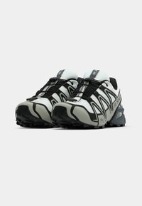 Sapatos de corrida de trilha com parte superior em malha e sintético nas cores branco e preto, com uma sola cinza texturada e sistema de atacadores elásticos.