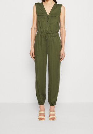 Vrouw draagt een mouwloze jumpsuit in olijfgroen met zakken aan de voorkant, een getailleerde taille, knopen aan de voorkant, elastische manchetten en beige open-toe sandalen met hak.