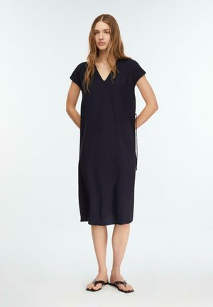 Kobieta stoi, ubrana w luźną, czarną sukienkę do kolan z krótkimi rękawami i czarne sandały na jednolitym białym tle.