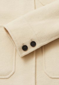 Manica di cappotto in lana color crema con texture morbida, caratterizzata da due bottoni neri vicino al polsino e dettagli di cuciture visibili.