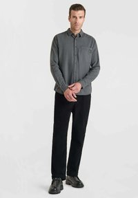 Camisa de manga larga gris con diseño, cierre de botones y bolsillo frontal, combinada con pantalones negros y zapatos negros, de pie frente a un fondo claro.