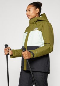 Žalia ir juoda izoliuota striukė su kapišonu, su balta krūtinės dalimi, užtrauktuku ir krūtinės kišene, nešiojama laikant trekking'o lazdas.