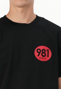 Tricou din bumbac negru cu un logo rotund roșu care conține "981" și "REPLAY". Guler rotund și mâneci scurte, textura netedă.