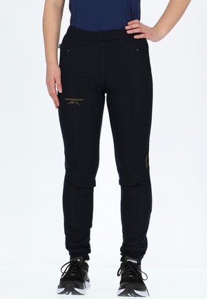 Vrouw die zwarte Swedemount sportbroek en zwarte Hoka hardloopschoenen draagt, staand met één hand op haar heup en de andere ontspannen.
