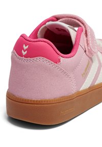 Baskets en daim rose avec un design à bande blanche et une bride à velcro. La semelle extérieure est en caoutchouc marron avec une finition texturée. Logo de la marque visible.