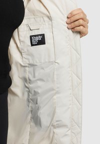 Ljust färgad kviltad jacka insida med en svart etikett som läser "Khujo original för ett fåtal." Slät tygtextur, ingen ytterligare hårdvara synlig.