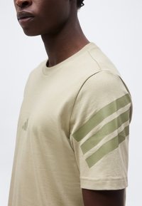 Άτομο που φοράει μπεζ μπλουζάκι με τρεις ελιά πράσινες ρίγες στο μανίκι και αντίστοιχο μικρό λογότυπο στο στήθος, κομμένο στο λαιμό.