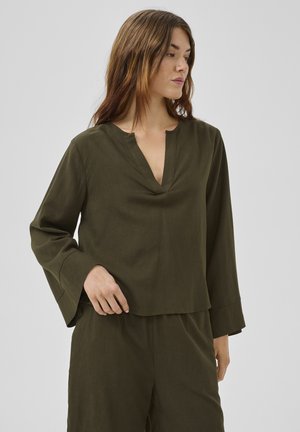 Frau trägt ein locker sitzendes, langärmeliges olivgrünes Oberteil und passende Hose, schaut seitlich vor einem einfarbigen hellen Hintergrund.